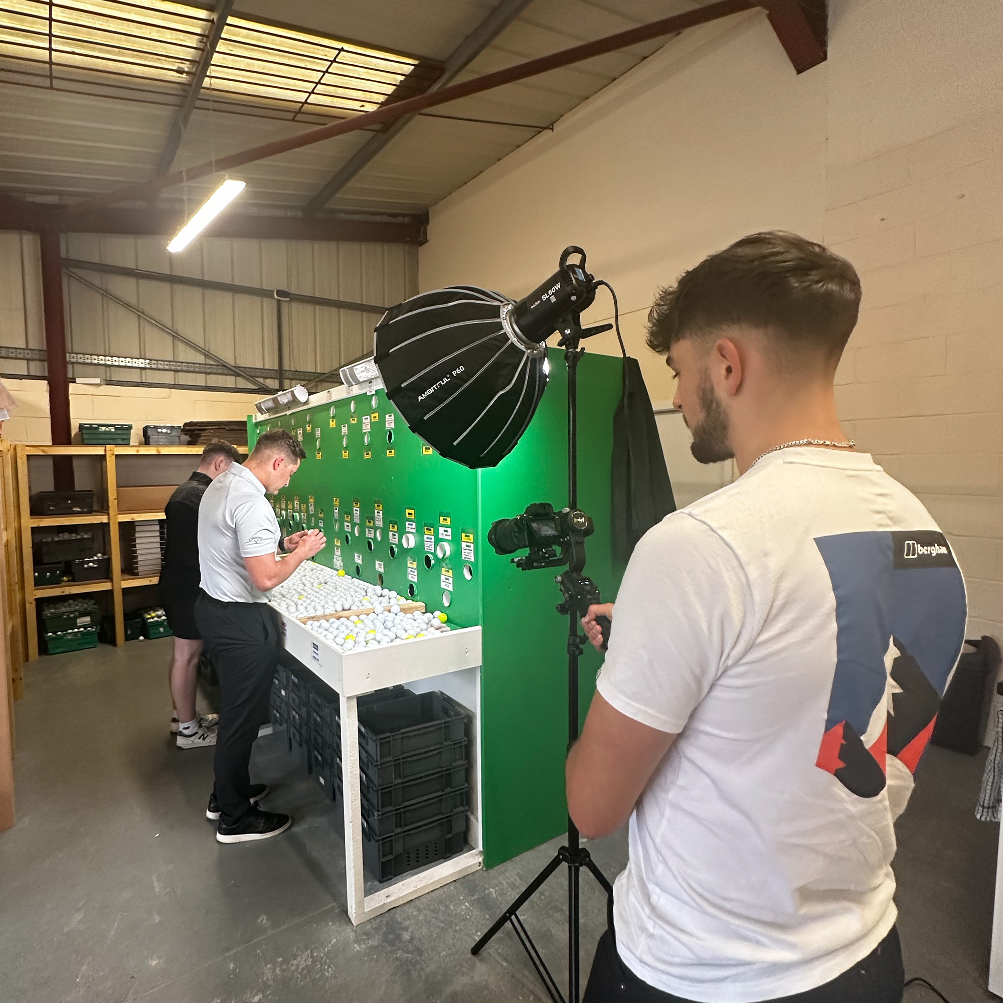 A videographer filming a man sorting golf balls in a workshop, with a softbox light on a stand